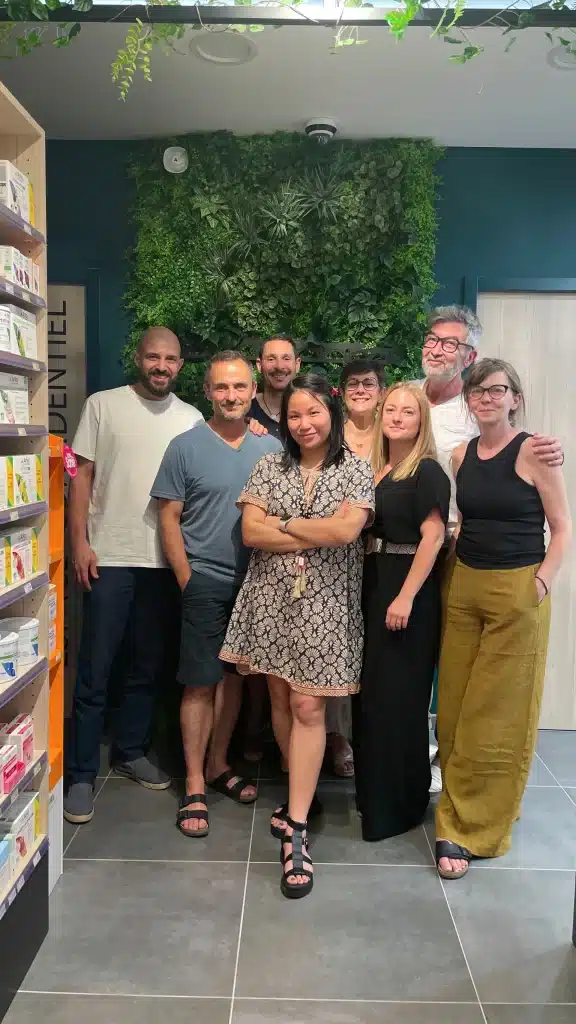 Un groupe de huit adhérents de Saint Laurent Presqu'île se tient devant un mur végétal intérieur, à côté d'une étagère de produits pharmaceutique.