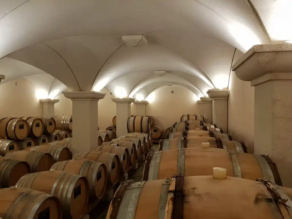 Intérieur d'une cave à vin voûtée avec de nombreuses rangées de fûts en chêne empilés, éclairés par des appliques murales encastrées.
