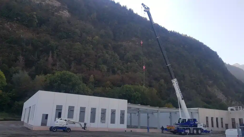 Chantier de construction en montagne avec une grue imposante, des éléments de bâtiment préfabriqués et une nacelle sur le côté.