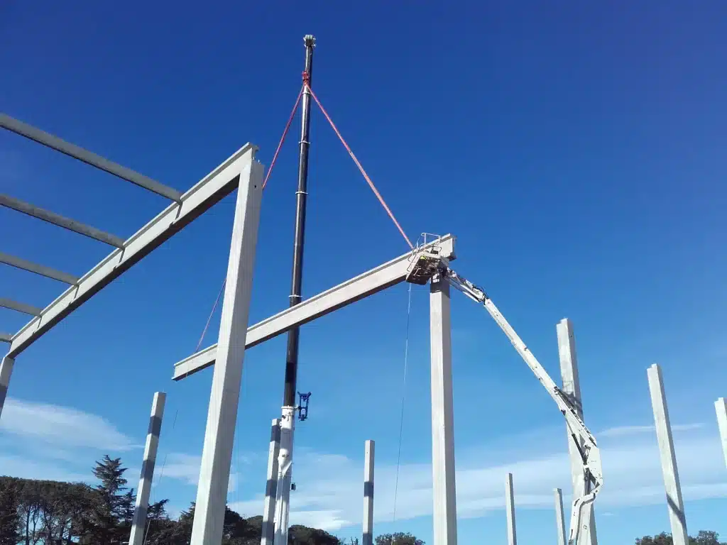 Une poutre en béton est soulevée par une grue sur un chantier de construction, avec une nacelle aérienne en soutien.