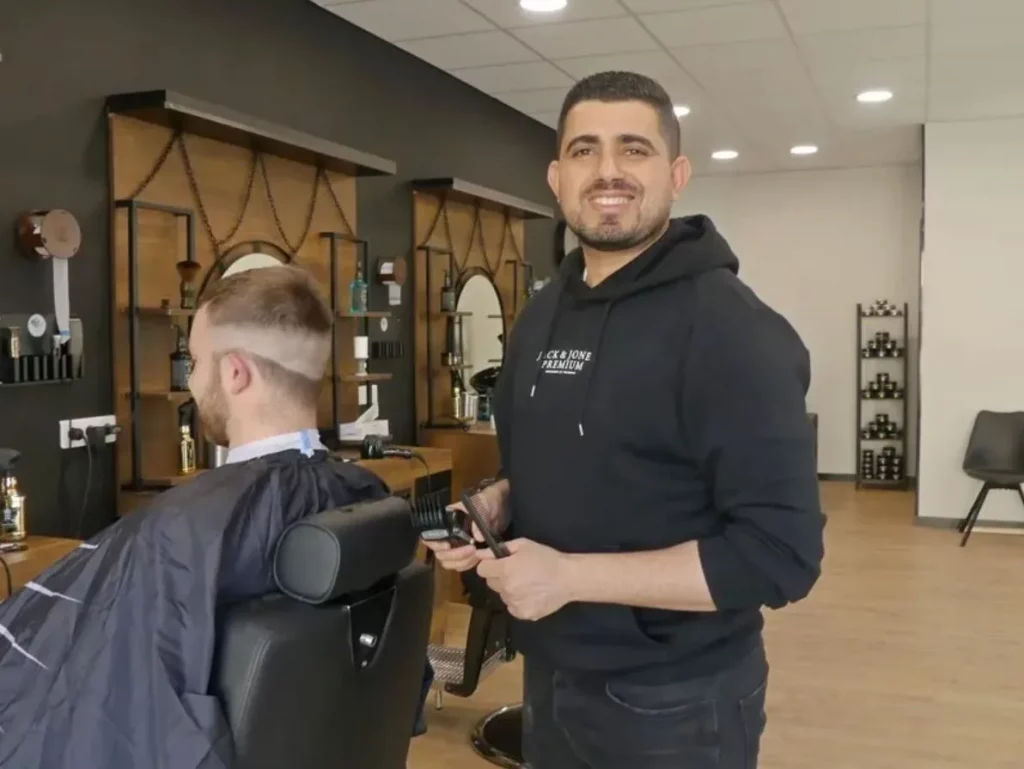 Coiffeur tenant des ciseaux et un peigne, souriant à la caméra pendant qu'il coupe les cheveux d'un client assis dans un fauteuil.