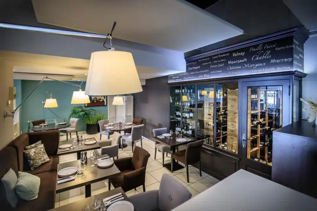 Ambiance chaleureuse d'un restaurant avec un coin salon cosy, des tables dressées et une grande cave à vin vitrée affichant diverses bouteilles et crus célèbres.
