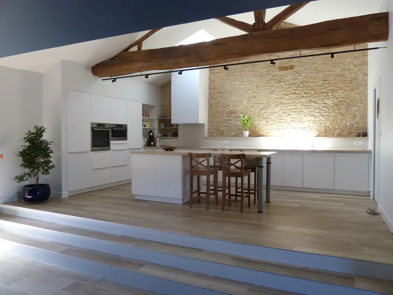 Cuisine moderne avec îlot central, meubles blancs, murs en pierre et poutres apparentes.