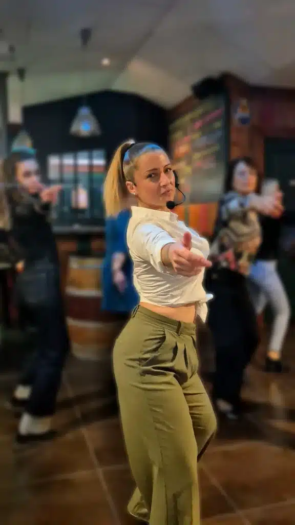 Une jeune femme danseuse au chignon blond, porte un micro et un chemisier blanc noué, avec un pantalon vert. Elle pointe son doigt vers l'avant.