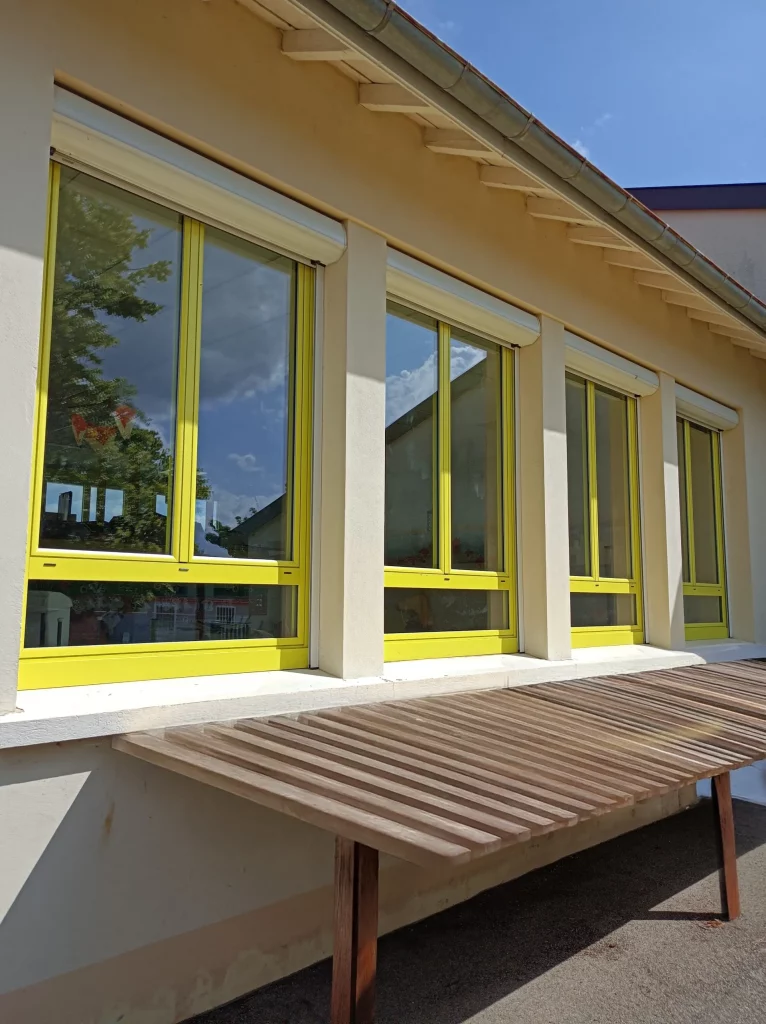 Façade d'un bâtiment avec plusieurs grandes fenêtres aux encadrements jaunes vives et volets roulants blancs, devant une table en bois.
