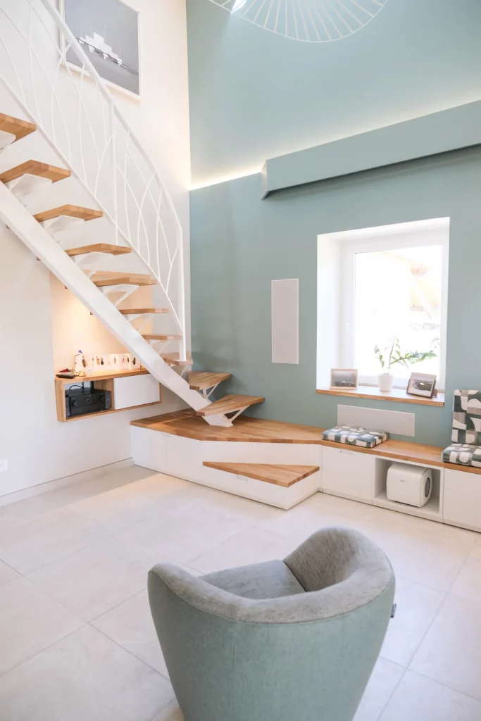 Escalier moderne blanc avec marches en bois, banquette intégrée avec rangements, fauteuil design et mur bleu pastel dans un salon.