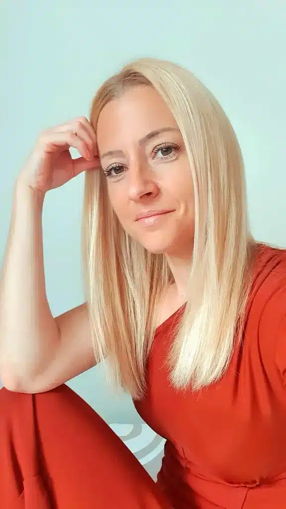 Portrait d'une femme aux cheveux blonds, regardant légèrement sur le côté, avec une main appuyée sur sa tête. Elle porte une tenue rouge.