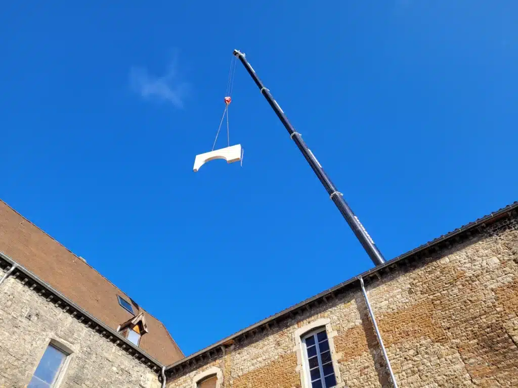 Une grue soulève un élément architectural blanc en forme d'arche au-dessus d'un bâtiment en pierre, sous un ciel bleu dégagé.