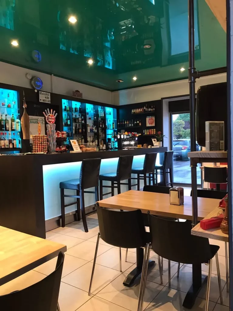 Ambiance intérieure d'un bar moderne avec un long comptoir noir rétroéclairé en bleu, des tabourets hauts et des tables dressées pour les clients.