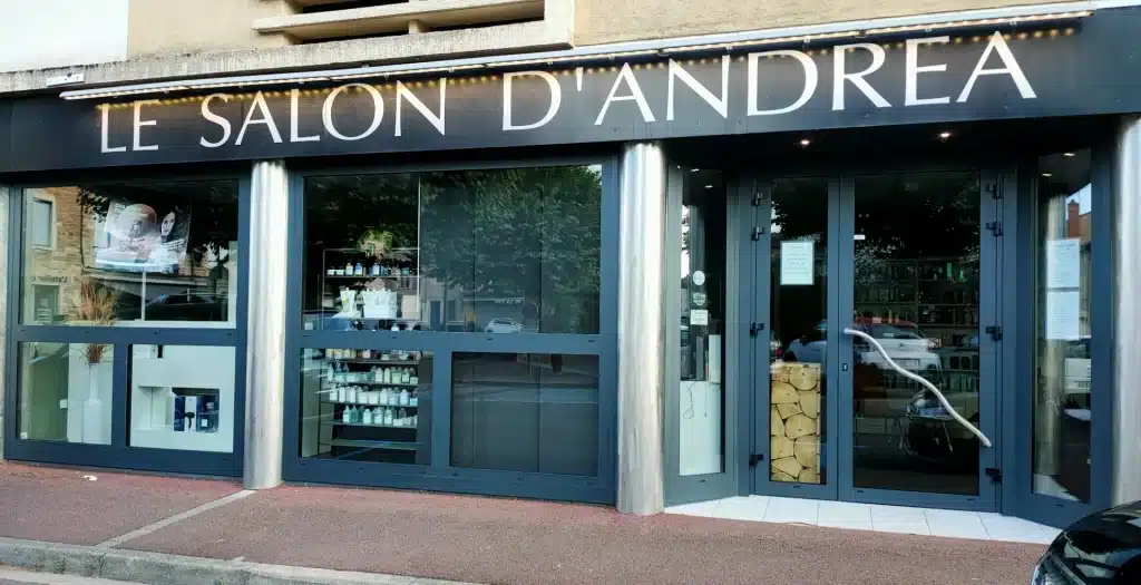 Façade du salon de coiffure "Le Salon D'Andrea" avec une grande vitrine, des portes vitrées et des présentoirs de produits.