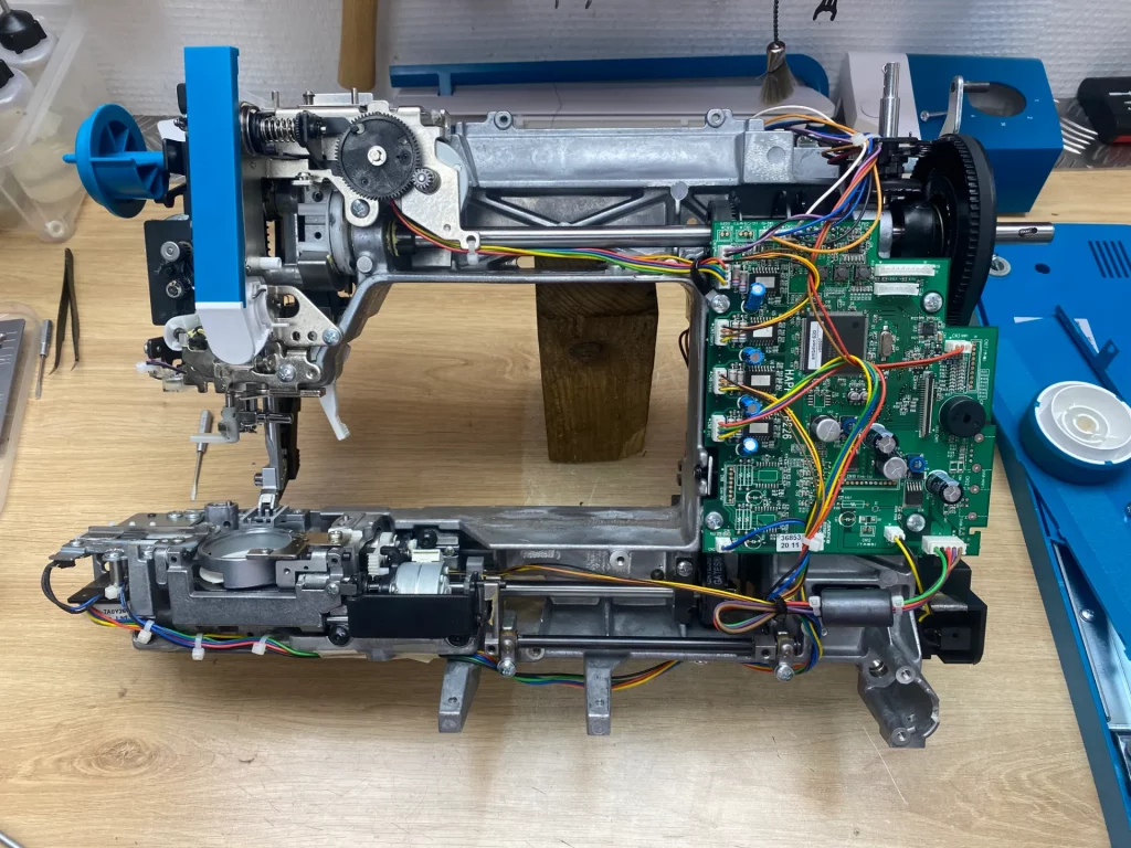 Machine à coudre démontée, montrant sa mécanique interne, ses fils et sa carte électronique verte avec des composants.