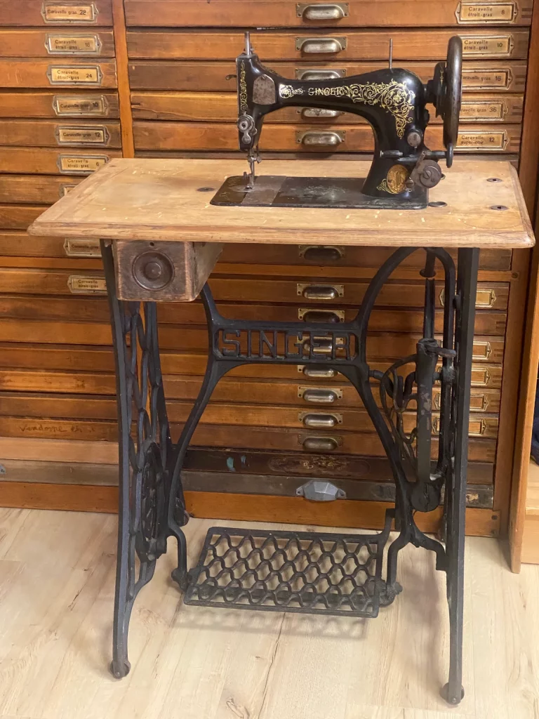 Machine à coudre Singer ancienne et décorée, posée sur un plateau en bois au-dessus d'un piedement en fer forgé avec pédale.