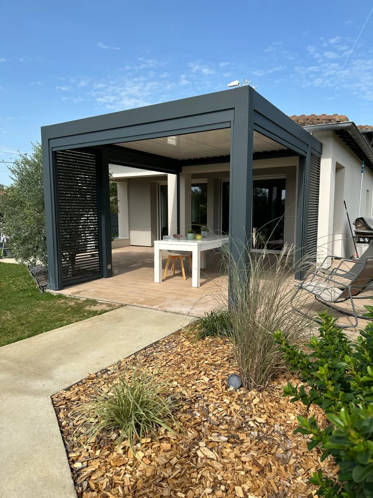 Table blanche et chaises sous une pergola moderne adossée à une maison, avec une terrasse en bois et un jardin aménagé.