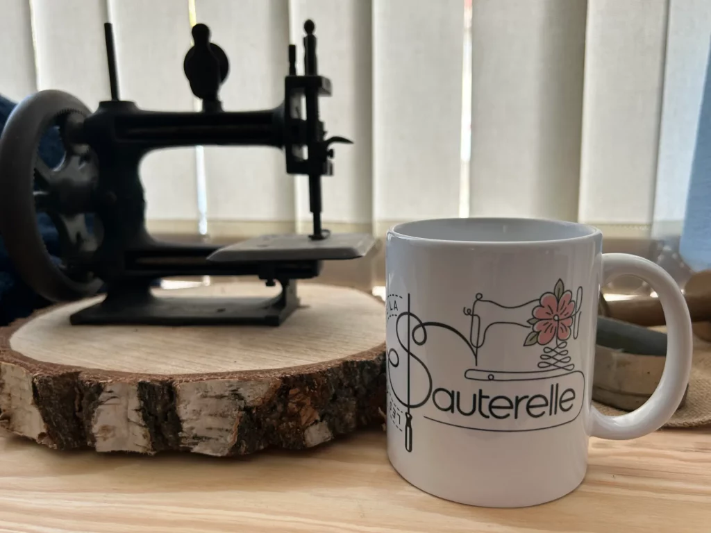Photographie d'une tasse à thé blanche décorée d'une machine à coudre et d'une fleur rose. Une petite machine à coudre ancienne est posée sur une tranche de bois à côté de la tasse.