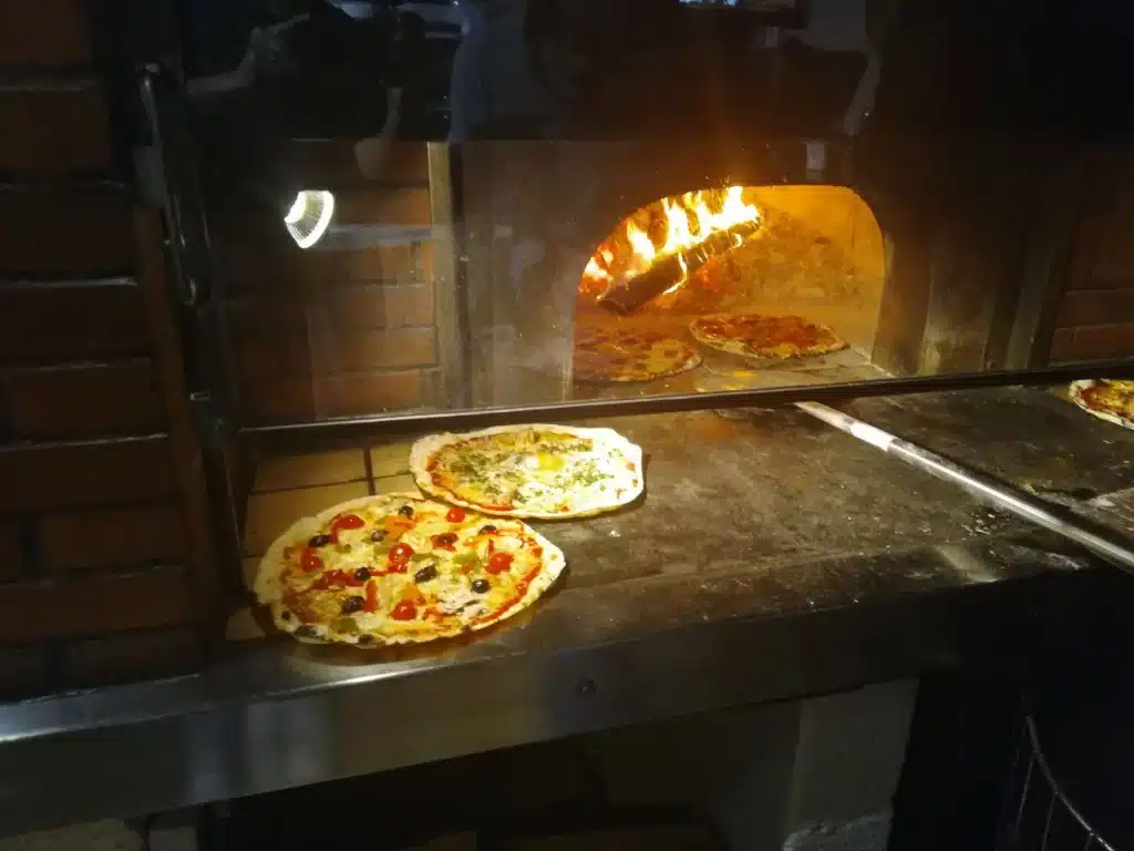 Plusieurs pizzas garnies, dont une aux olives et tomates cerises, reposent sur un plan de travail devant un four à bois ardent.