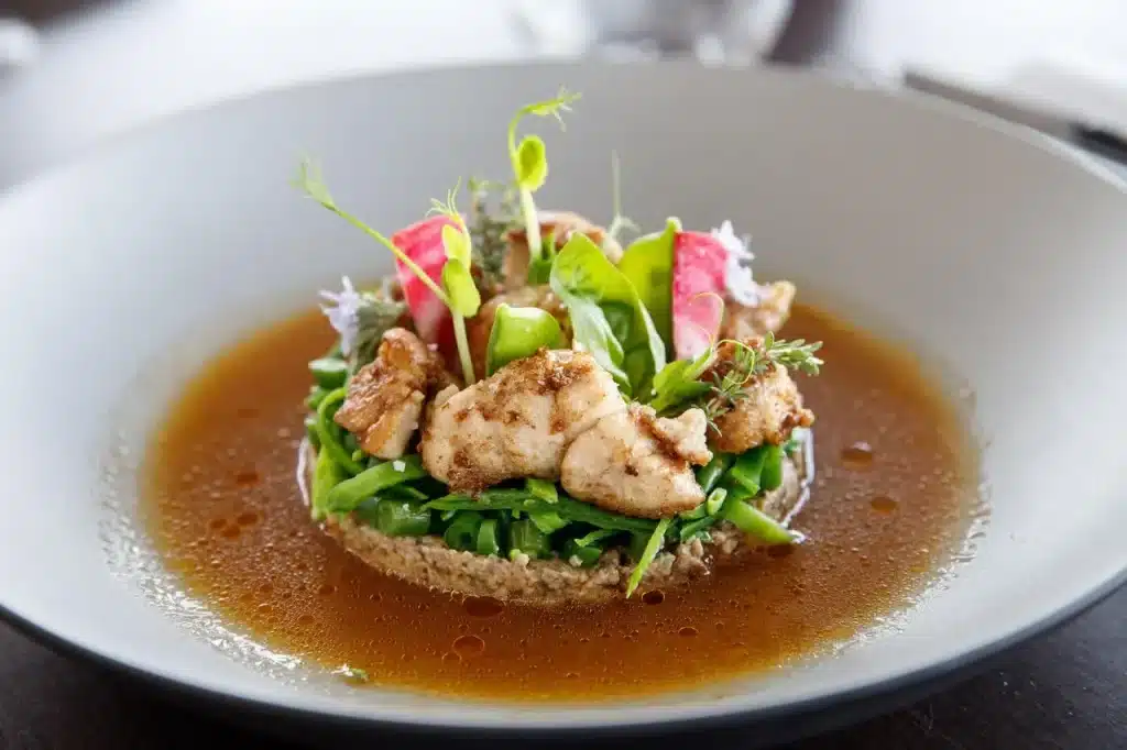Gros plan d'un plat de poulet servi sur des légumes verts et du grain, baignant dans un bouillon clair, décoré de radis et de pousses tendres.