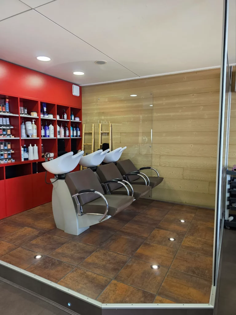 Banquette de fauteuils de coiffure marrons avec bacs de lavage blancs intégrés, devant une étagère rouge remplie de produits capillaires.