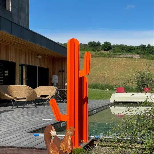 Sculpture de cactus orange vif sur une terrasse en bois, avec des chaises en rotin, une piscine et des collines en arrière-plan.