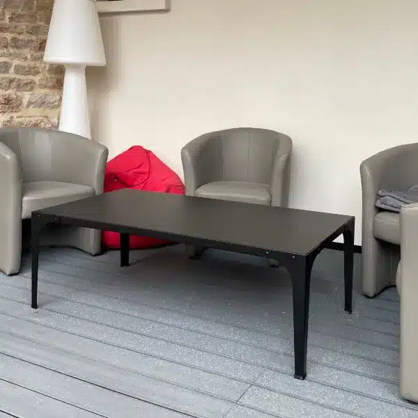 Table basse rectangulaire grise avec pieds noirs, entourée de fauteuils gris et d'un pouf rouge, sur un sol type terrasse.