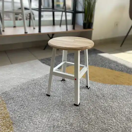Un tabouret blanc à pieds métalliques et une assise ronde en bois clair, posé sur un tapis gris et ocre.