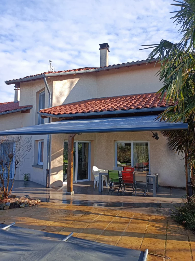 Vue d'une terrasse de maison avec une table et des chaises colorées sous un auvent, devant un jardin et une piscine.