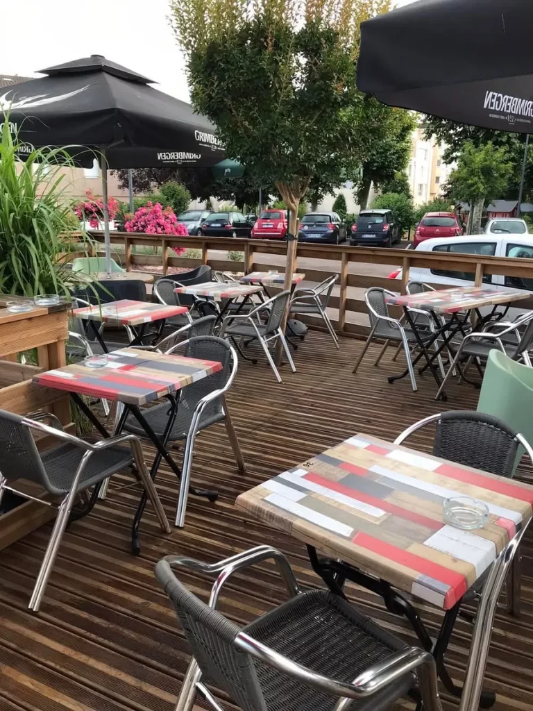 Terrasse extérieure d'un restaurant avec plusieurs tables vides et leurs chaises grises, sous des parasols noirs.