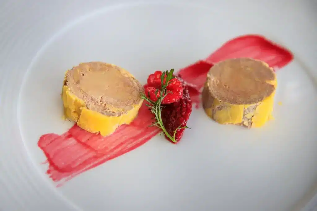 Deux tranches de foie gras servies avec une purée de framboises et des framboises fraîches sur une assiette blanche.