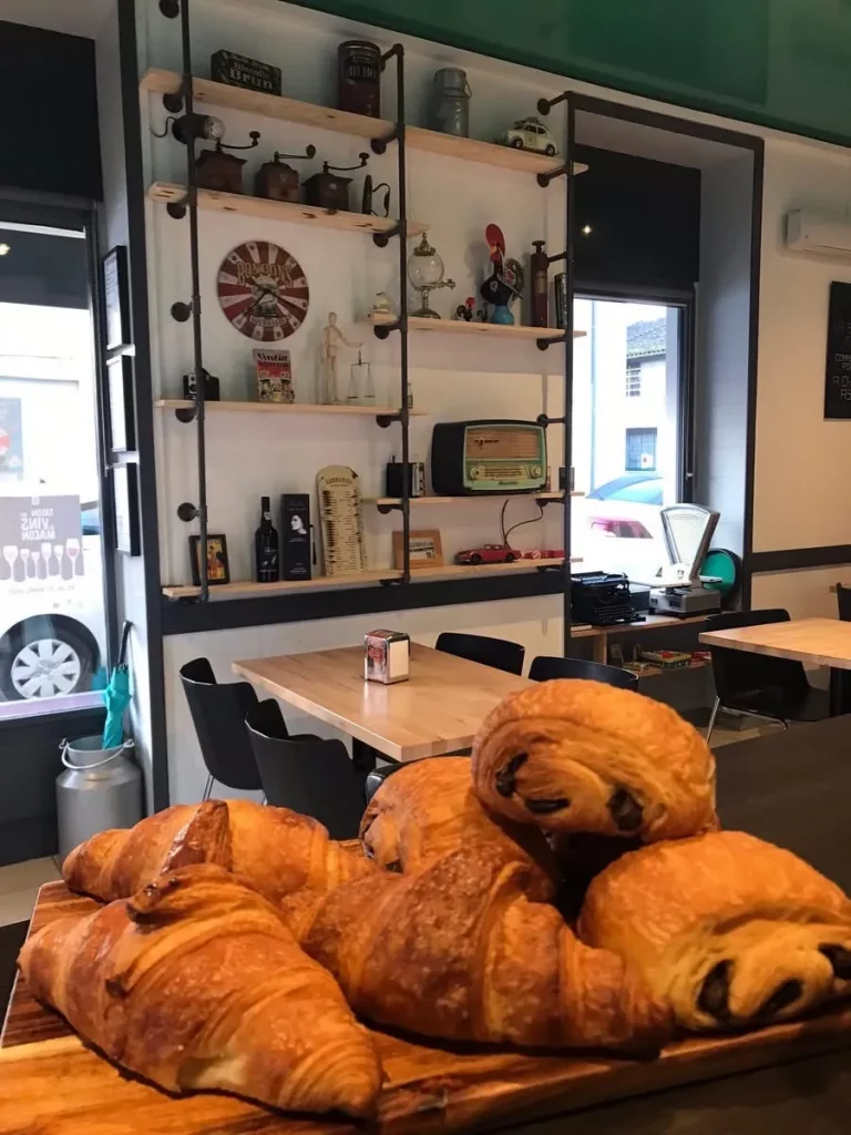 Gros plan sur des viennoiseries, dont des croissants et des pains au chocolat, disposées sur un plateau en bois. En arrière-plan, une étagère murale remplie d'objets décoratifs et une table avec des chaises.